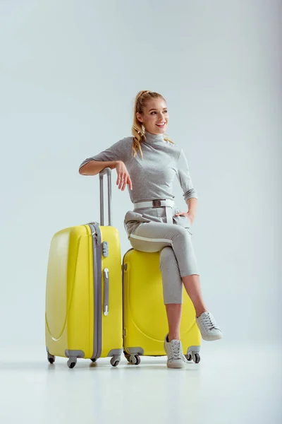 Donna sorridente seduta su valigie con gambe incrociate su sfondo grigio, concetto di viaggio — Stock Photo