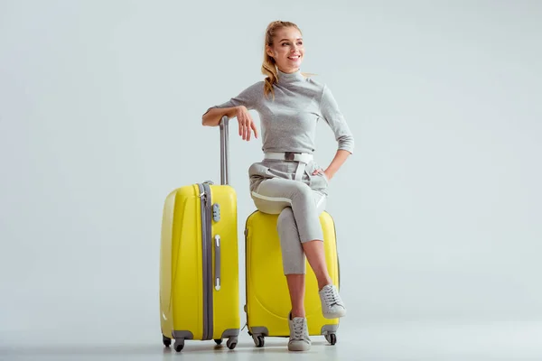 Belle femme souriante assise sur des valises aux jambes croisées sur fond gris, concept de voyage — Photo de stock