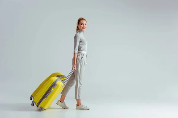 Жінка, що несе жовті валізи і дивиться на камеру на сірому фоні, концепція подорожей — стокове фото