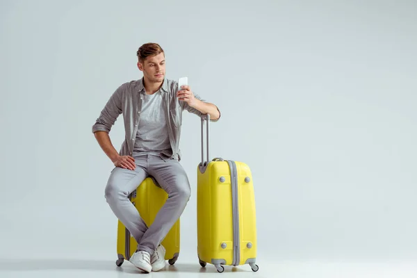 Hombre guapo sentado en la maleta amarilla y el uso de teléfono inteligente sobre fondo gris, concepto de viaje - foto de stock