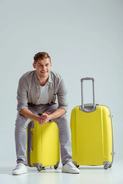 Hombre guapo sonriente sentado en una maleta amarilla con las manos dobladas sobre fondo gris, concepto de viaje - foto de stock