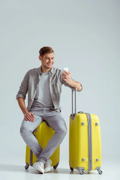 Улыбающийся мужчина, сидящий на желтом чемодане и использующий смартфон на сером фоне, концепция путешествия — стоковое фото