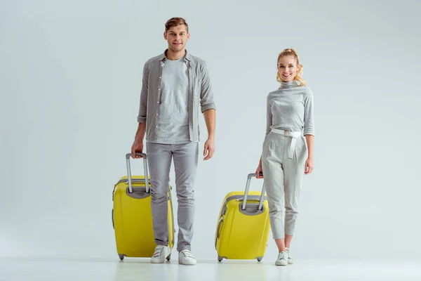 Улыбающаяся пара с желтыми чемоданами и смотрящая на камеру на сером фоне, концепция путешествия — стоковое фото