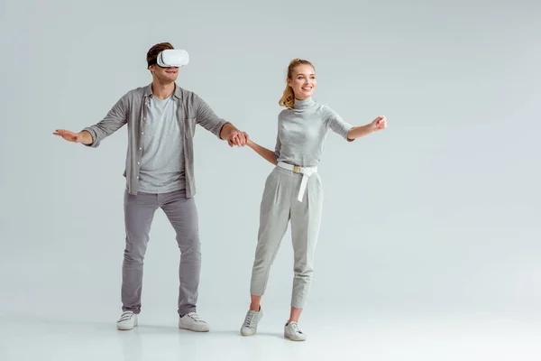 Glückliches Paar Händchen haltend, während Mann virtuelle Realität auf grauem Hintergrund erlebt — Stockfoto