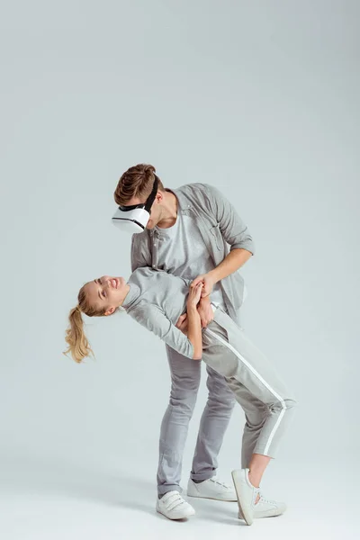 Чоловік тримає жінку в обіймах, маючи досвід віртуальної реальності на сірому фоні — стокове фото