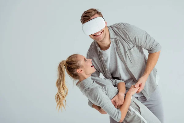 Hombre sosteniendo sonriente mujer en abrazo mientras tiene experiencia de realidad virtual aislado en gris - foto de stock