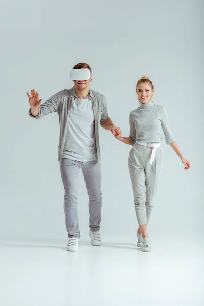 Sonriente pareja cogida de la mano mientras el hombre experimenta realidad virtual sobre fondo gris - foto de stock