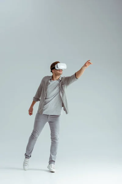 Чоловік у сірому одязі жестикулює в гарнітурі віртуальної реальності на сірому фоні — стокове фото