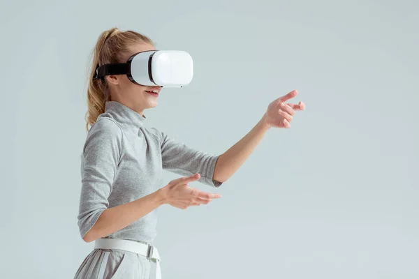 Mujer sorprendida haciendo gestos mientras tiene experiencia de realidad virtual aislada en gris - foto de stock