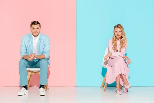 Couple with folded hands sitting on chairs and looking at camera on pink and blue background — Stock Photo