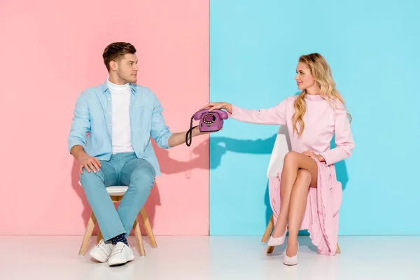 Couple sitting on chairs and holding purple vintage telephone on pink and blue background — Stock Photo