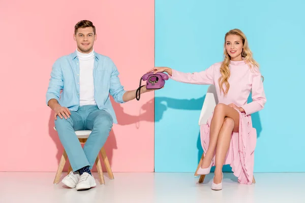 Beautiful couple sitting on chairs and holding purple vintage telephone on pink and blue background — Stock Photo