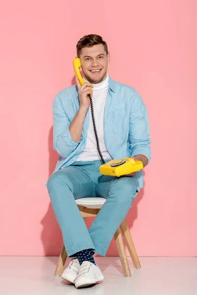 Усміхнений чоловік сидить і розмовляє на жовтому старовинному телефоні з рожевим фоном — стокове фото