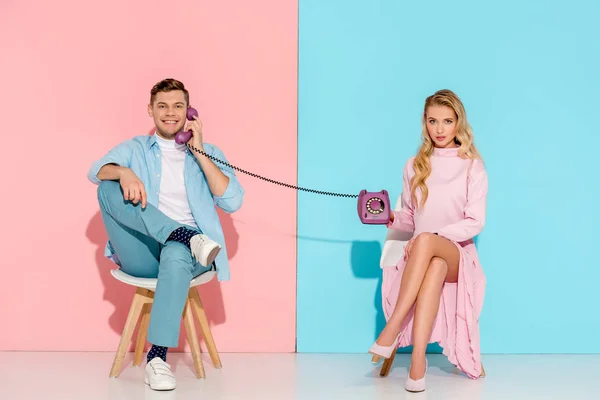 Dissatisfied woman holding purple vintage telephone while handsome man talking on pink and blue background — Stock Photo