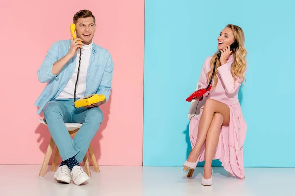 Feliz pareja sentada y conversando en teléfonos antiguos con fondo rosa y azul - foto de stock