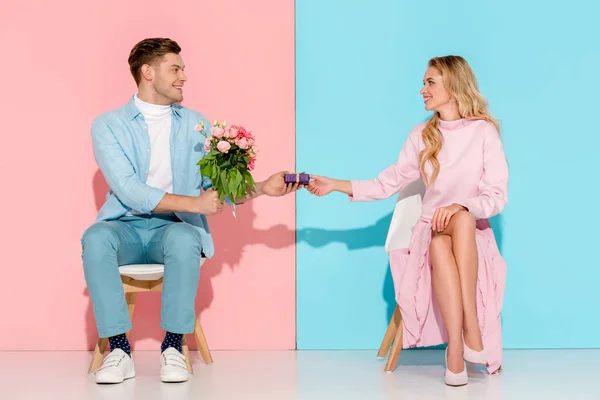 Belo casal sentado em cadeiras enquanto homem apresentando caixa de presente com buquê de flores para mulher no fundo rosa e azul — Fotografia de Stock