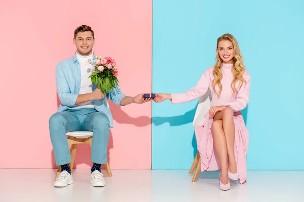 Pareja sentada en sillas mientras el hombre presenta caja de regalo con ramo de flores a la mujer sonriente sobre fondo rosa y azul - foto de stock