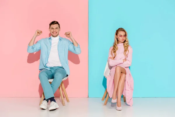 Dissatisfied woman with arms crossed and cheering man with clenched fists sitting on chairs on pink and blue background — Stock Photo