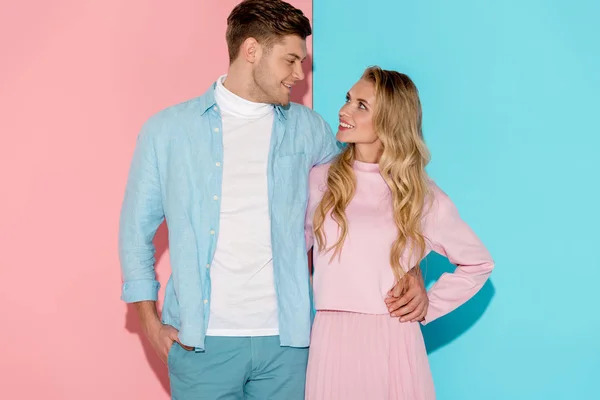 Lächelnder Mann und Frau, die sich umarmen und einander auf rosa und blauem Hintergrund anschauen — Stockfoto