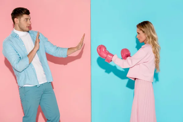 Frightened man gesturing with hands near aggressive woman in pink boxing gloves on pink and blue background — Stock Photo