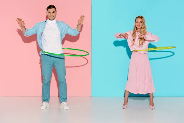 Beautiful couple looking at camera and exercising with hula hoops on pink and blue background — Stock Photo