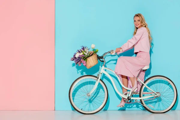 Smiling woman in pink clothing riding bicycle with flower basket on pink and blue background — Stock Photo