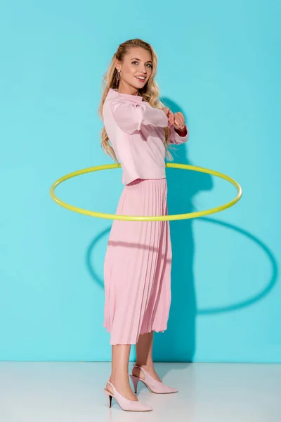 Schöne Frau in pinkfarbener Kleidung, die in die Kamera schaut und mit Hula-Hoop-Reifen auf türkisfarbenem Hintergrund trainiert — Stockfoto