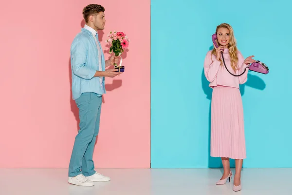 Мужчина с цветочным букетом и подарочной коробкой, улыбающаяся женщина разговаривает по старинному телефону с розовым и голубым фоном — Stock Photo