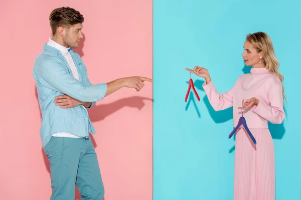 Handsome man pointing with finger at woman and choosing empty clothes hangers on pink and blue background — Stock Photo