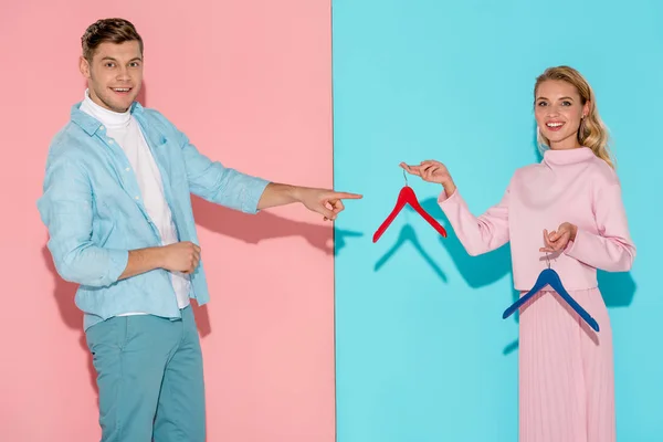 Smiling man pointing with finger at woman and choosing empty clothes hangers on pink and blue background — Stock Photo