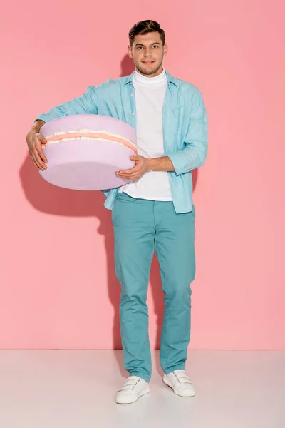 Man in blue clothing holding big macaroon model and looking at camera on pink background — Stock Photo