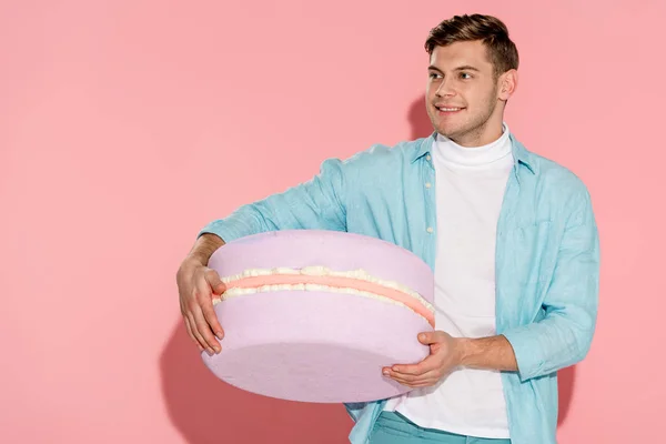 Handsome man in blue clothing holding big macaroon model on pink background — Stock Photo