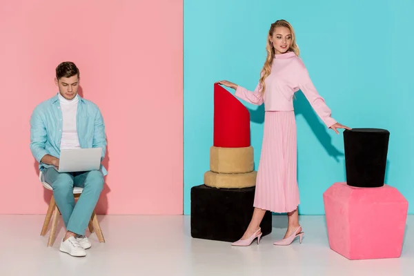 Beautiful woman posing with nail polish and lipstick models while man sitting on chair and using laptop on pink and blue background — Stock Photo