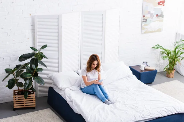 High angle view of woman with ginger hair resting in bed and using smartphone at home on weekend — Stock Photo