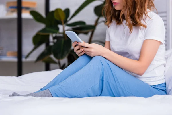 Imagen recortada de la mujer con el pelo de jengibre descansando en la cama y el uso de teléfono inteligente en casa en fin de semana - foto de stock