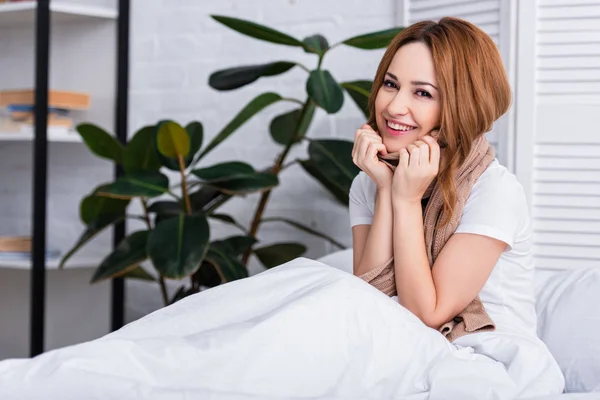 Mujer enferma sonriente con bufanda sentada en la cama en casa y mirando a la cámara - foto de stock