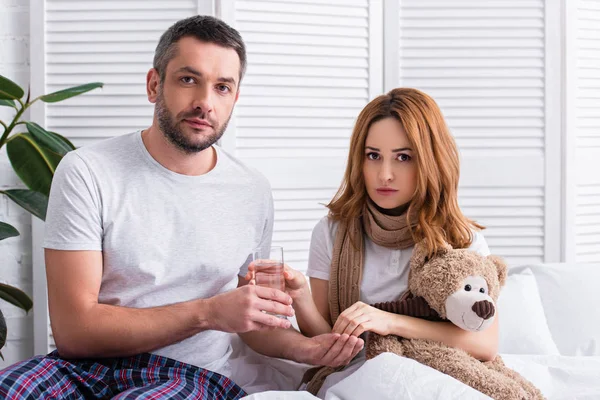 Marido dando pastillas y vaso de agua a la esposa enferma en el dormitorio, mirando a la cámara - foto de stock