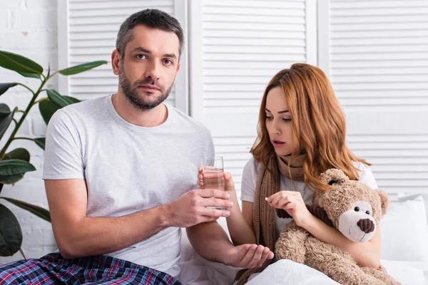 Marito che dà pillole e bicchiere d'acqua alla moglie malata con orsacchiotto in camera da letto — Foto stock