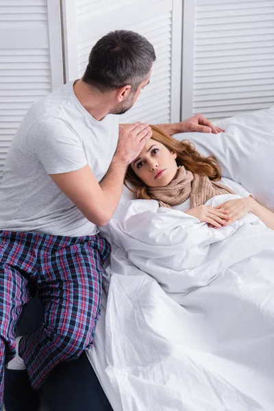Marido tocando la frente de la esposa enferma con fiebre y comprobando la temperatura en la cama - foto de stock