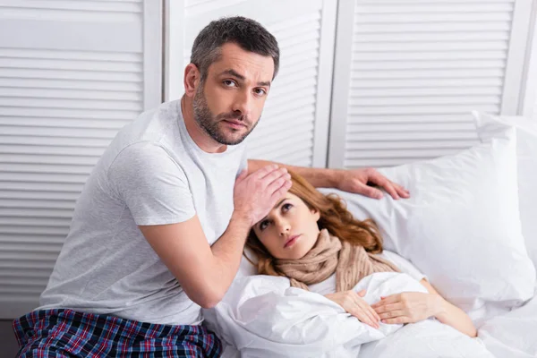 Bonito marido tocando testa de doente esposa com febre na cama, olhando para a câmera — Fotografia de Stock