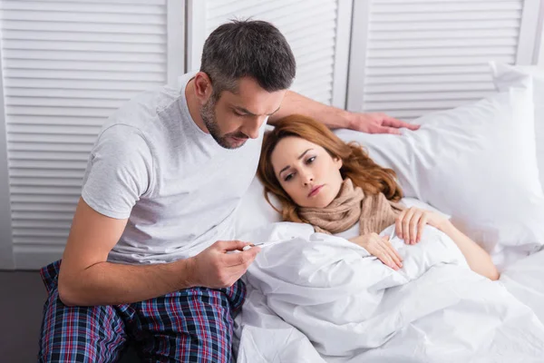Муж проверяет температуру больной жены и смотрит на термометр в спальне — стоковое фото