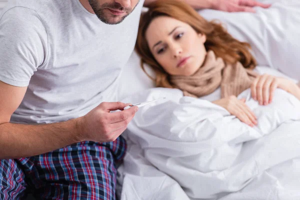 Cropped image of husband checking temperature of sick wife and holding thermometer in bedroom — Stock Photo