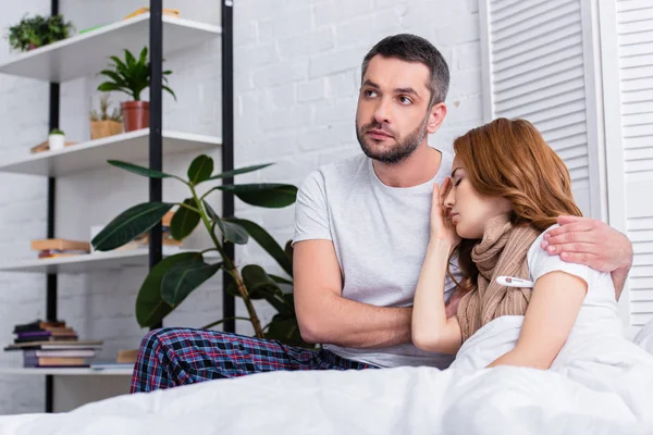 Красивый муж обнимает больную жену в спальне и смотрит в сторону — стоковое фото