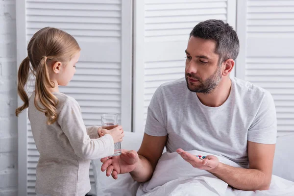Daughter giving glass of water to father to take pills in bedroom — Stock Photo