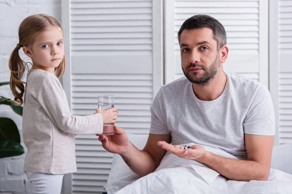 Дочь дает стакан воды папе, чтобы принять таблетки в спальне, глядя в камеру — стоковое фото