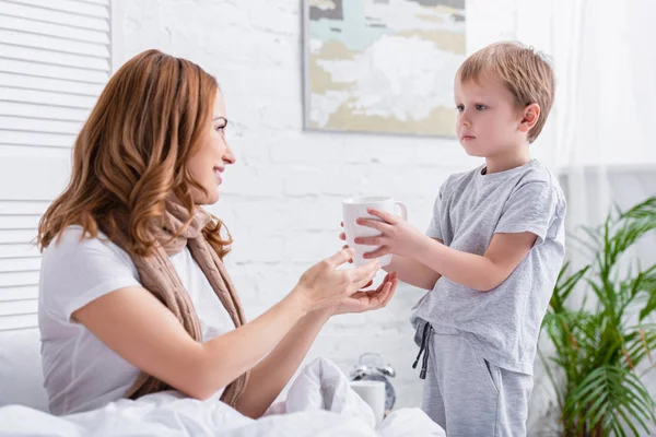 Visão lateral do filho dando xícara de chá para a mãe doente no quarto — Fotografia de Stock