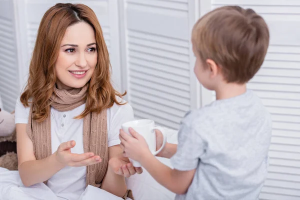 Son giving cup of tea to sick happy mother in bedroom — Stock Photo