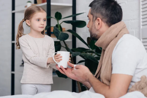 Adorable daughter giving cup of tea to sick dad in bedroom — Stock Photo