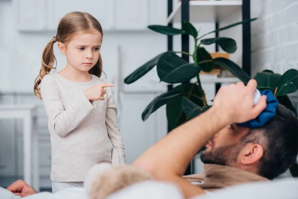 Adorable daughter pointing on sick father touching head with ice pack in bedroom — Stock Photo