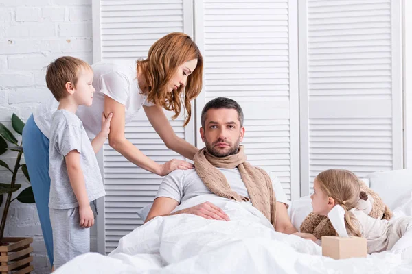 Femme et enfants prenant soin de père malade dans la chambre, il couché sur le lit et regardant la caméra — Photo de stock